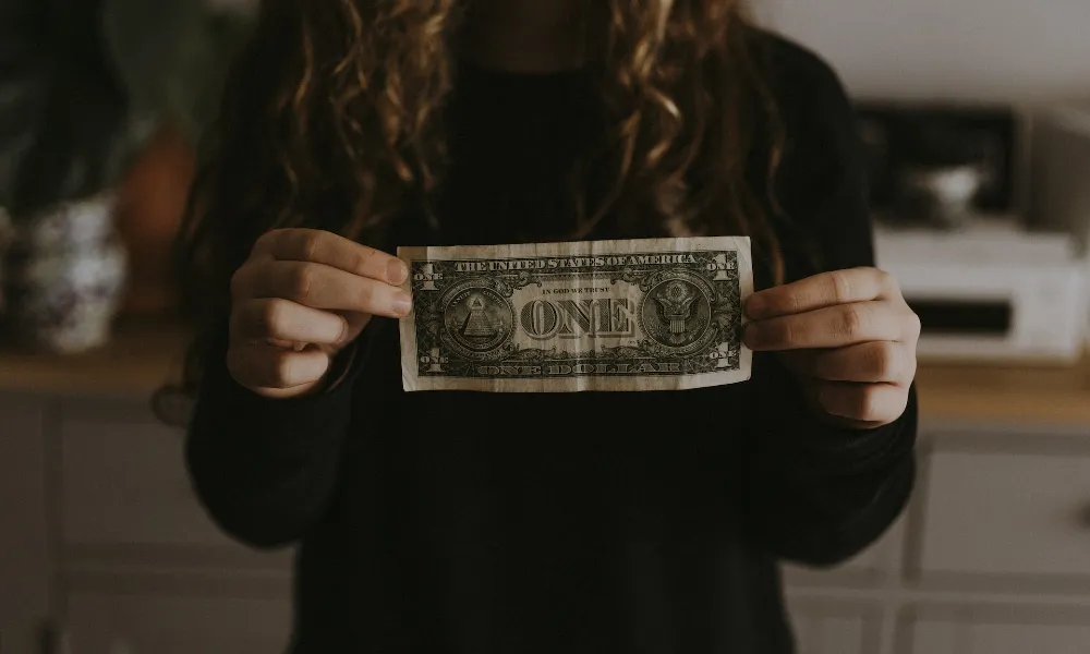 A person with long, brown curly hair wearing black holding out a one dollar bill, photo by Annie Spratt on Unsplash